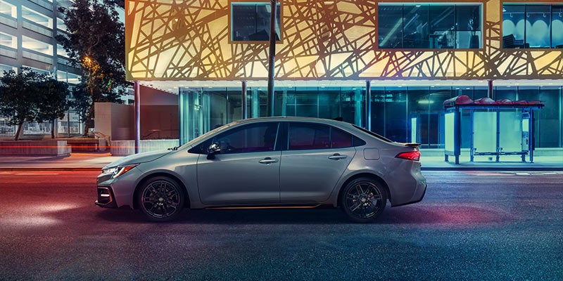 A grey Toyota Corolla drives down a city street at night.