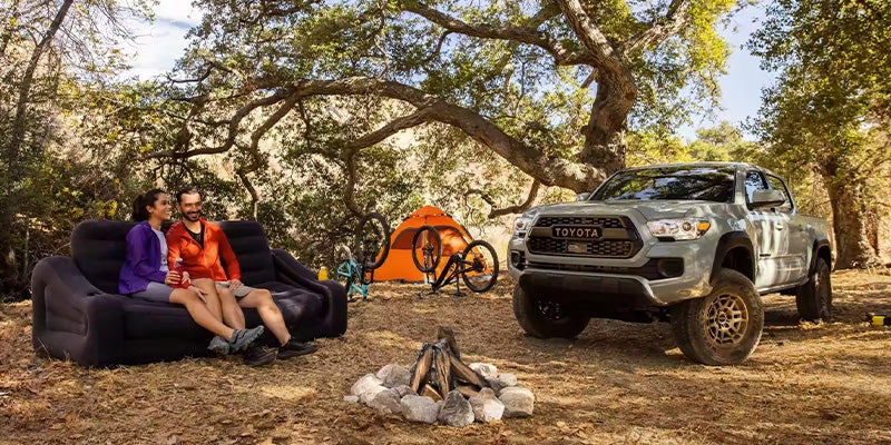 A couple sits at a campsite with a grey Toyota Tacoma parked under the trees.