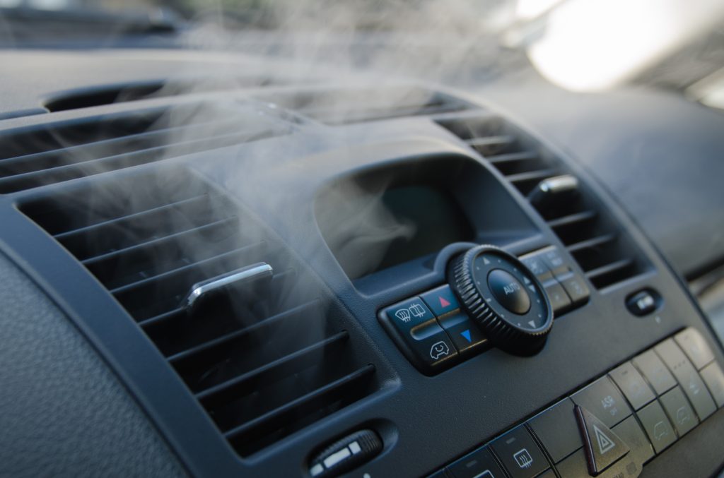 Close-up of a car dashboard showing air vents emitting vapor, with climate control buttons and a hazard light button visible.
